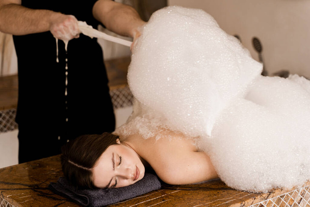 Chica atractiva relajarse en el spa. Masajista está haciendo pelado de espuma en hammam spa turco - Foto, Imagen