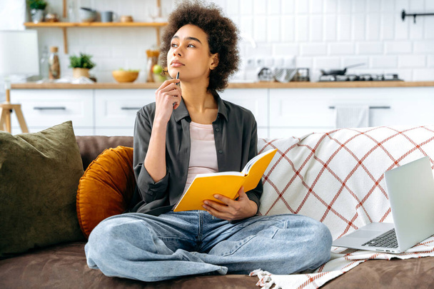 Thoughtfully stylishly dressed curly haired african american young woman, student or freelancer, sitting in the living room on the sofa with laptop and notebook, study homework, looking away, thinking - Fotografie, Obrázek