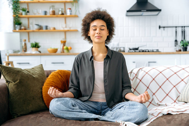 Relaxation and meditation concept. Happy peaceful relaxed african american woman in casual clothes, sitting alone at home in living room on the couch and meditating in the lotus position, smiling - Foto, afbeelding