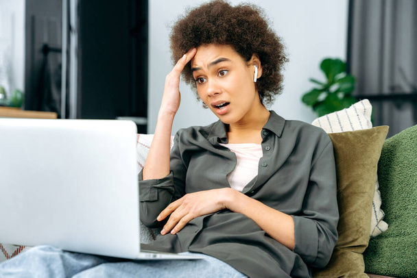 Потрясенная ошеломленная африканская женщина с короткими вьющимися волосами, в стильной одежде, сидит на диване в гостиной с ноутбуком, работает онлайн или проводит досуг в Интернете, смотрит в изумлении на ноутбук - Фото, изображение
