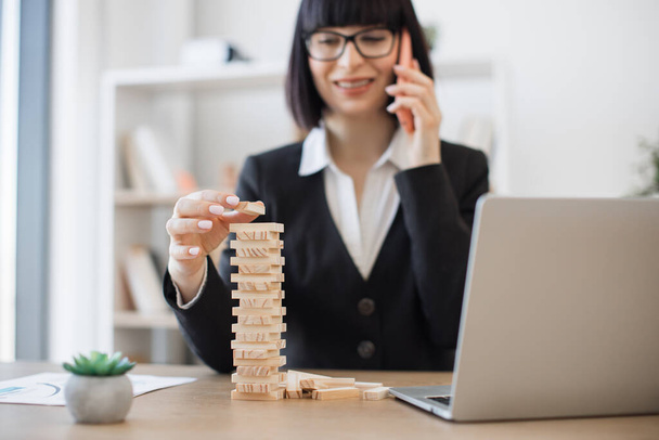 Young caucasian businesswoman in glasses holding wooden block of jenga game while speaking on smartphone on office background. Creative manager training control risk by building tower at work. - Photo, image