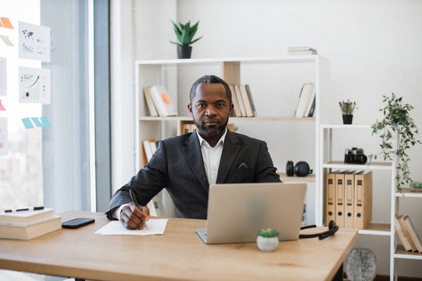 Confident office administrator in smart suit sitting at writing desk with modern gadgets. Focused multicultural white-collar worker writing down information from laptop screen at office. - Фото, зображення