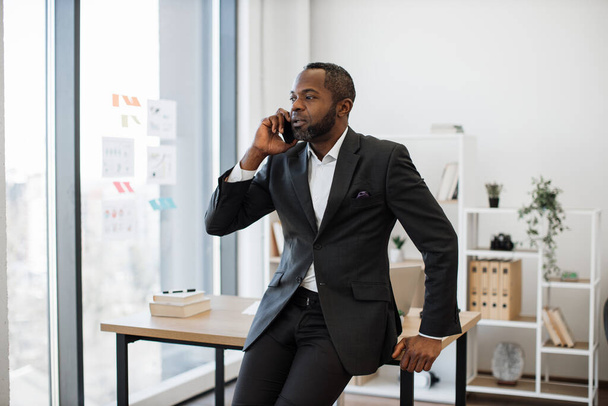 Confident african american businessman in formal attire speaking on cell phone while leaning against office desk in airy workplace. Project manager discussing new program with colleagues in office. - Фото, изображение