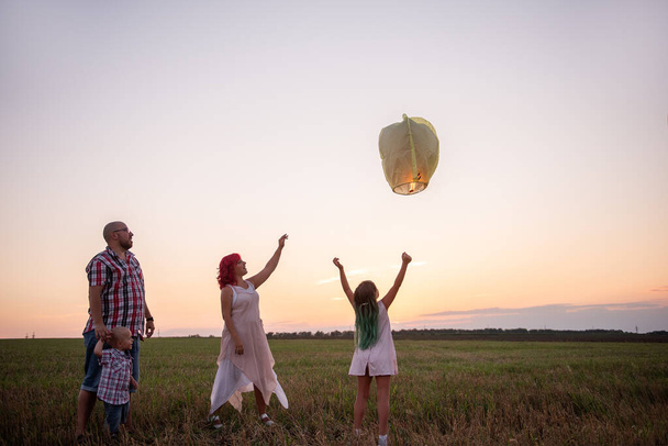 Diversity family launch yellow sky lantern in field at sunset. Mother, father holding Chinese paper lantern, daughter, son wave hands. Making wishes for holidays. Concept of hope. Teamwork - Фото, изображение