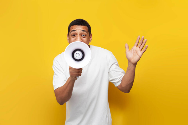 young African American guy in white T-shirt announces the news through loudspeaker on yellow isolated background, man shouts into megaphone - Zdjęcie, obraz