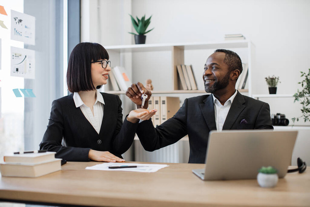 Joyful multiracial businessman in formal attire giving house keys to his caucasian female coworker while sitting behind writing desk in office. Real estate professionals finding buyers perfect home. - Фото, зображення