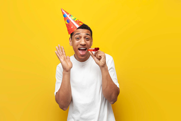 joyful african american guy in festive hat celebrates his birthday and rejoices on yellow isolated background - Fotó, kép