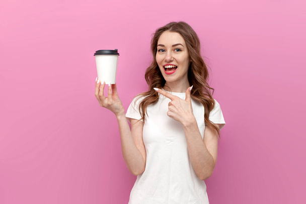 young cute woman with paper cup of coffee on pink isolated background, girl in white t-shirt shows and advertises drink - Fotó, kép