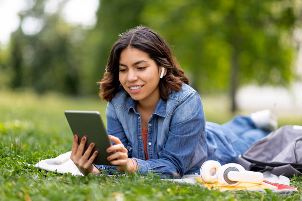 Linda jovem árabe feminino descansando ao ar livre com tablet digital, sorrindo Millennial mulher do Oriente Médio usando gadget moderno enquanto deitado no gramado no parque, vestindo fones de ouvido sem fio, espaço de cópia - Foto, Imagem