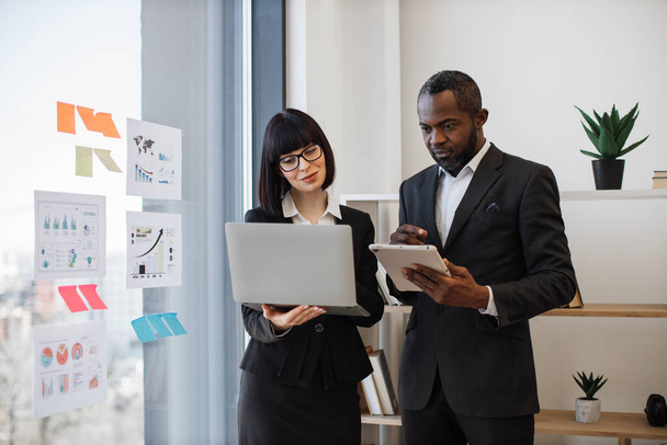 Caucasian woman and multiracial man in business clothes holding electronic devices while communicating in modern workplace. Professional coworkers being involved in teamwork using laptop and tablet. - Foto, Bild