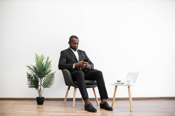 Hombre de negocios afroamericano en traje inteligente escribiendo en el teléfono celular mientras está sentado cerca de la mesa de café con el ordenador portátil en el interior minimalista. Relajado gerente charlando con los clientes a través de Internet. - Foto, Imagen