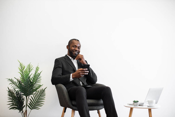 African man in business suit smiling cheerfully on camera while using modern smartphone at office center. Laptop and coffee cup on table near. Concept of people, business and technology. - Valokuva, kuva