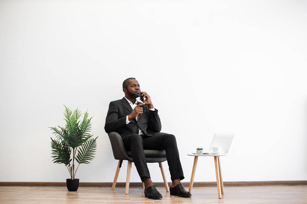 オフィスセンターで働いている間、ビジネスでアフリカ人男性が現代のスマートフォンで話しているのを確信しています。遠隔地のパートナーとの作業上の問題を解決する集中雇用主. - 写真・画像