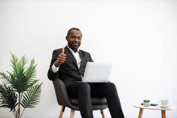 Diligent african employee in formal wear smiling sincerely at camera while typing on wireless laptop at office with modern minimalist design showing thumb up. People, work and technology concept. - Foto, immagini