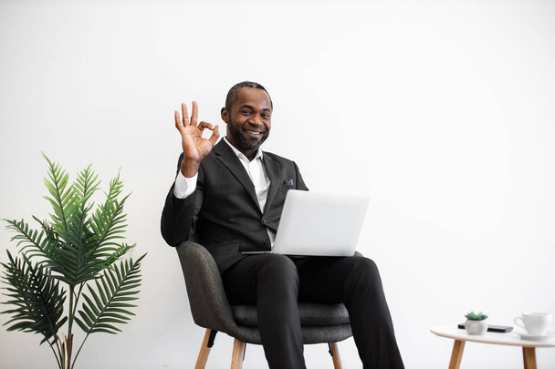 Diligent african employee in formal wear smiling sincerely at camera while typing on wireless laptop at office with modern minimalist design showing sign ok. People, work and technology concept. - Foto, immagini
