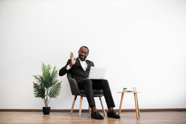 Confused african man in black suit having video conference on wireless laptop while working at office. Intense conversation with coworkers on distance. Modern lifestyles of business people. - Foto, immagini