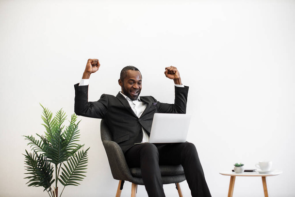 African american young businessman dressed in formal suit gesturing emotionally from happiness while looking on computer screen at office. Success in business, luck and human emotions concept. - Фото, изображение