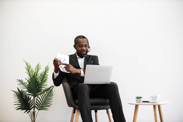 Competent african american financier presenting charts and graphs during online video meeting on modern laptop. Formally dressed man in headset sitting at office and having meeting on distance. - Photo, Image