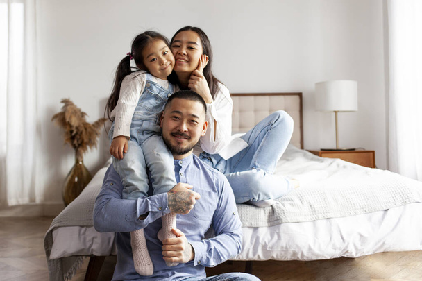 glückliche asiatische Familie mit kleiner Tochter sitzt zu Hause in der Nähe des Bettes und lächelt, koreanisches Mädchen sitzt auf Papas Armen und umarmt Mama, elterliche Liebe und Fürsorge - Foto, Bild