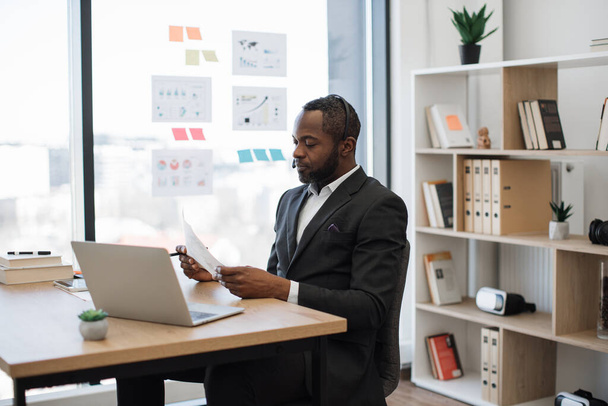 Formally dressed male entrepreneur sitting at workspace and having online meeting with colleagues on laptop. African american man in headset examining some documents holding in hands. - Foto, afbeelding