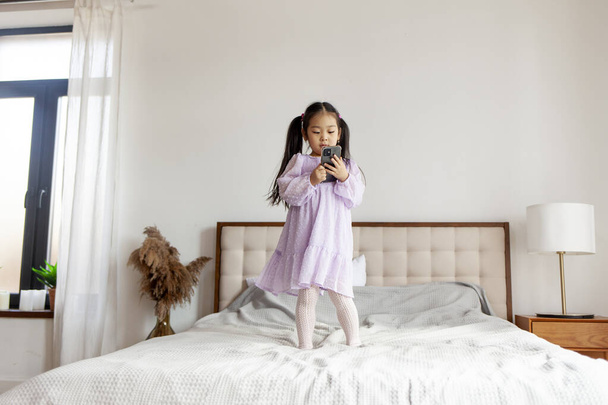 menina asiática pouco pulando na cama em casa e usando smartphone, a criança olha para o telefone em vestido roxo e danças - Foto, Imagem