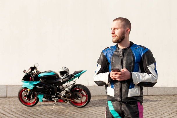 gars motocycliste en équipement de protection professionnel se tient près de la moto de sport et utilise smartphone contre mur blanc, coureur de moto professionnel tenant le téléphone et regardant de côté - Photo, image