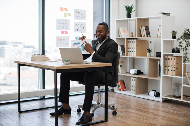 Homem africano sorridente em roupa formal sentado na mesa do escritório e tendo videoreunião. Funcionário masculino usando fone de ouvido sem fio e laptop para conferência on-line com colegas. - Foto, Imagem