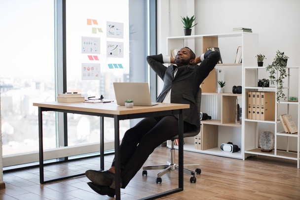 Office worker in black suit holding hands behind head and keeping eyes closed while sitting at workplace with laptop on table. Tired african american man having rest during coffee break at work. - 写真・画像