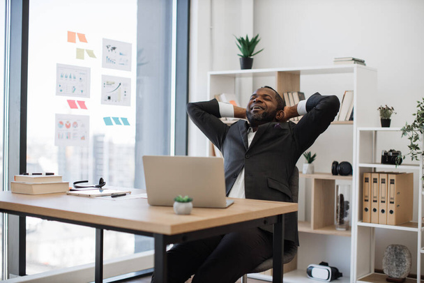 Fatigued and sleepy african man in suit yawning in chair while overwhelmed with work at office. Laptop with documents lying on table. People, technology and overwork concept. - 写真・画像