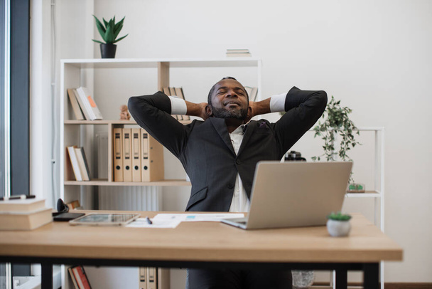 African american male employee sitting comfy in chair with hands behind head while taking break from wireless laptop at modern office. Concept of exhaustion, workspace and deadline. - Фото, зображення