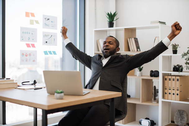 Sleepy and tired african american man in formal wear yawning and stretching arms after hard working hours on wireless laptop at office. Concept of people, technology and overwork. - Photo, Image