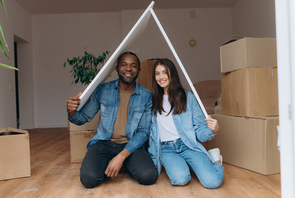 una pareja se muda a su propia casa, juntos hacen un gesto del techo de la casa, se sientan en el suelo entre cajas de cartón - Foto, imagen