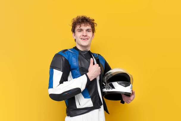 レザージャケットの若い男のオートバイは、黄色の隔離された背景にヘルメットと笑顔を保持しています,保護具のプロのオートバイの肖像画 - 写真・画像