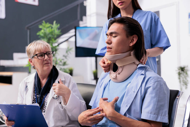 Medical staff helping asian patient with neck brace removing cervical neck collar after injury accident during consultation.. Injured young adult healing fracture and trauma in hospital waiting area - Fotografie, Obrázek