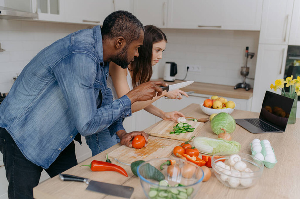 Una pareja casada cocina juntos usando un ordenador portátil, buscando recetas en Internet. Un hombre afroamericano y su esposa preparan una ensalada en la cocina - Foto, Imagen