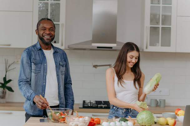 Un couple interracial heureux qui prépare à manger dans la cuisine. Un homme afro-américain et une femme blanche préparent une salade - Photo, image