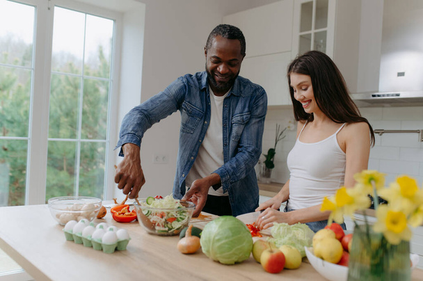 Una feliz pareja interracial preparando comida en la cocina. Hombre afroamericano y una mujer caucásica preparan una ensalada - Foto, imagen