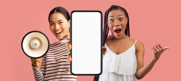 Offerta fantastica. Due donne multietniche che utilizzano megafono e mostrano grande smartphone vuoto con schermo bianco, donne multiculturali che fanno annuncio con altoparlante e pubblicità nuova app, Mockup - Foto, immagini