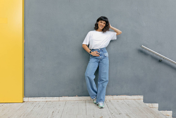 Foto de cuerpo entero de una mujer elegante y guapa con un peinado corto que usa una camiseta blanca y jeans que sostiene la mano y sonríe al aire libre sobre una pared gris. - Foto, imagen