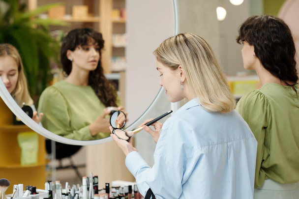 Młody klient ze szczotką i kompaktowym proszkowym testowaniem produktów kosmetycznych stojąc przed lustrem w kosmetycznym supermarkecie - Zdjęcie, obraz