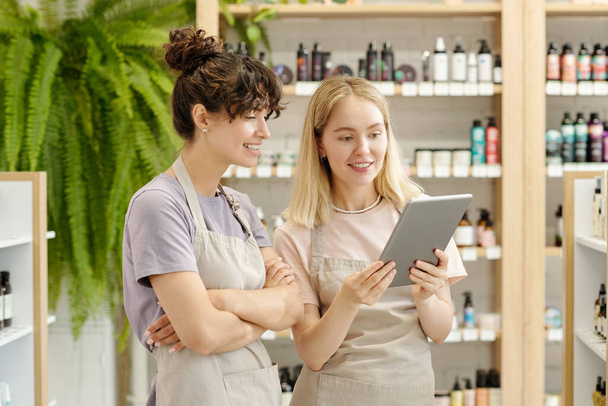 Zwei junge lächelnde Verkäuferinnen in Arbeitskleidung blicken auf den Tablet-Bildschirm, während sie Sortimentsgespräche führen oder Kunden online beraten - Foto, Bild