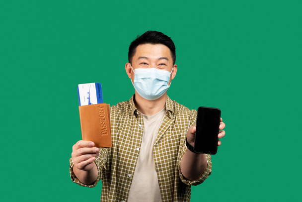 Koreaanse man van middelbare leeftijd in het gezicht masker met paspoort met tickets en toont mobiele telefoon met mockup voor app op groene achtergrond. Man reizend tijdens covid lockdown - Foto, afbeelding