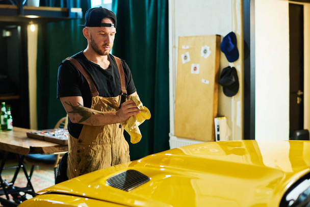 Young repairman in workwear rubbing dirty hands with duster over hood of yellow electric car while standing in front of it in garage - Φωτογραφία, εικόνα