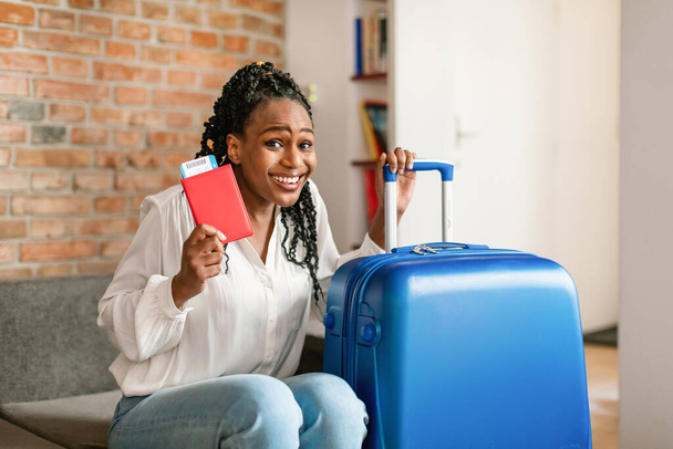 Mujer negra alegre sentada al lado de la maleta brillante, con pasaporte internacional con entradas y sonriendo a la cámara, lista para el viaje. Concepto de viaje de verano - Foto, imagen