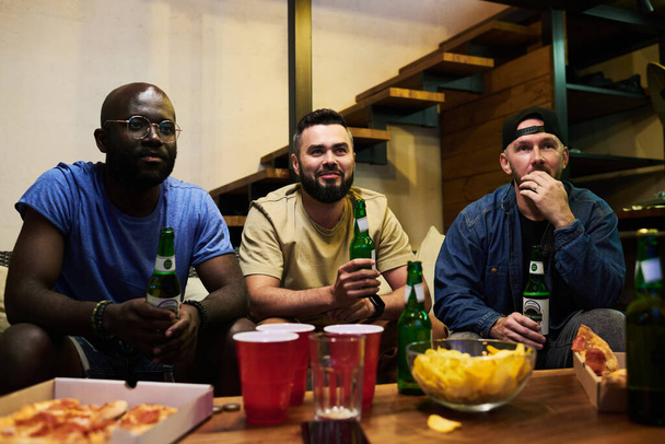 Drie interculturele mannelijke voetbalfans verzameld door tafel met bier en snack in de voorkant van de tv in de garage om te kijken naar uitzending van de wedstrijd - Foto, afbeelding