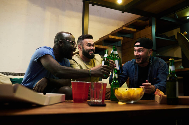 Młodzi, weseli, międzyrasowi kumple i fani piłki nożnej brzęczący butelkami piwa nad stołem z kubkami i przekąskami podczas świętowania zwycięstwa - Zdjęcie, obraz