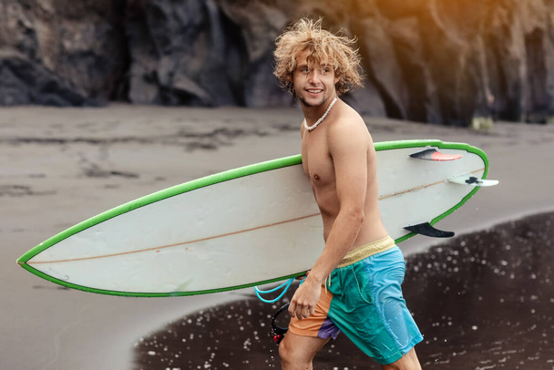 Kıvırcık sarı saçlı, sörf tahtası olan genç sörfçü adam okyanusun yanından geçiyor. Ekstrem su sporları yapıyor, sörf yapıyor. Seyahat ve sağlıklı yaşam tarzı kavramı. Spor gezisi hedefi - Fotoğraf, Görsel