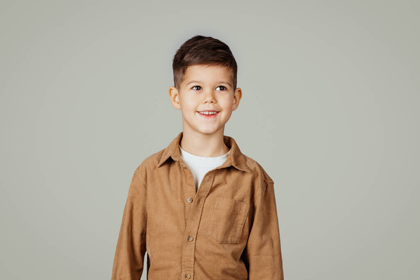 Smiling caucasian 6 years old little boy pupil in casual looks at empty space isolated at gray studio background. Lifestyle, childhood, education, study, advertising and offer - Photo, Image