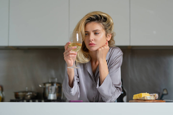 Housewife drink wine at kitchen. Domestic lifestyle. Woman holding a glass of wine in her kitchen. Blond beautiful girl with phone drinking wine. Morning of pretty woman using social media on phone - Photo, image