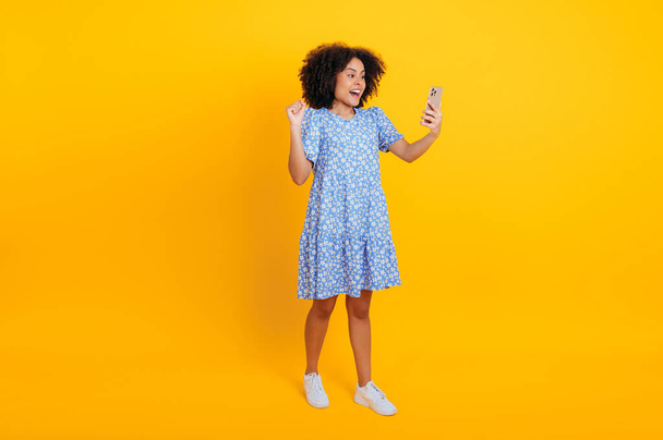 Femme bouclée africaine américaine ou brésilienne pleine longueur en robe d'été, tient le téléphone intelligent à la main, faisant un geste oui, se réjouissant du succès, de la victoire, des nouvelles, se tenir debout sur un fond orange isolé - Photo, image